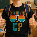 Garçon souriant en classe de CP avec son sac à dos, heureux de faire sa rentrée scolaire, portant un T-shirt humoristique « En route vers le CP ».