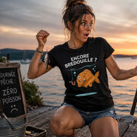 Femme pêcheuse surprise au bord d'un lac au coucher du soleil, tenant une canne à pêche avec une expression amusée après une journée sans réussite, portant un T-shirt humoristique sur la pêche.