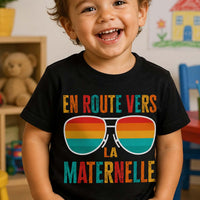 Jeune enfant de 2 à 3 ans souriant portant un T-shirt noir « En route vers la maternelle » dans une salle de classe colorée, idéal pour illustrer une tenue de première rentrée en école maternelle.