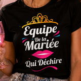 Mockup photoréaliste d'une femme portant un t-shirt noir avec le visuel « Équipe de la mariée qui déchire » lors d'un enterrement de vie de jeune fille (EVJF). Entourée de ses amies dans une ambiance festive avec décor rose, ballons, coupes de champagne et accessoires de fête, cette scène met en valeur un t-shirt humoristique idéal pour les demoiselles d'honneur et l'équipe de la mariée.