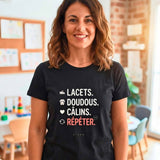 T-shirt noir de femme porté par une Atsem dans une classe de maternelle, avec le design "LACETS. DOUDOUUS. CÂLINS. RÉPÉTER." et les icônes associées, entourée de dessins d'enfants.