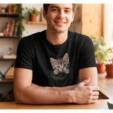 T-shirt noir unisexe en coton porté par un jeune homme dans un café lumineux, mettant en valeur l'illustration trompe-l'œil d'un chaton tigré sortant de la poche de poitrine