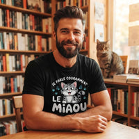 T-shirt noir pour homme en coton, porté par un homme barbu souriant dans une librairie chaleureuse, avec l'illustration d'un chaton gris et le texte blanc "JE PARLE COURAMMENT LE MIAOU" et des empreintes de pattes. Un chat tigré est assis sur une étagère en arrière-plan