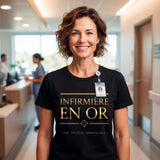 T-shirt noir pour femme en coton, porté par une infirmière souriante dans une unité de soins moderne. Le design "INFIRMIÈRE EN OR" avec le texte doré et la croix est clairement visible et centré