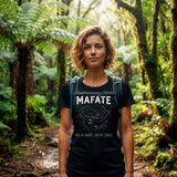 T-shirt noir pour femme en coton porté par une randonneuse marchant sur un sentier en sous-bois dense, de face, avec le visuel de carte et le texte "MAFATE PAS DE ROUTE, PAS DE STRESS" sur le devant