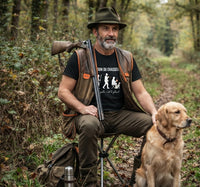 Un chasseur barbu en tenue traditionnelle (chapeau, gilet) assis dans les bois sur un tabouret pliant, tenant son fusil et caressant son chien golden retriever, portant le t-shirt 'L'ÉVOLUTION DU CHASSEUR'. Scène de chasse en forêt