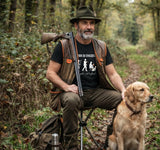 Un chasseur barbu en tenue traditionnelle (chapeau, gilet) assis dans les bois sur un tabouret pliant, tenant son fusil et caressant son chien golden retriever, portant le t-shirt 'L'ÉVOLUTION DU CHASSEUR'. Scène de chasse en forêt