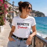 Femme portant un t-shirt blanc avec l'inscription « Bichette D'amour », souriante sur une promenade en bord de mer, avec l'océan, des fleurs et une ambiance estivale en arrière-plan.