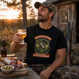 Homme portant un t-shirt noir « Chasseur d'Apéros » avec une illustration de saucissons, de verres et d'un tire-bouchon, assis devant un chalet en forêt lors d'un apéritif après une journée de chasse, avec une planche de charcuterie et une boisson à la main.