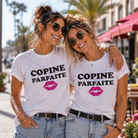 Deux meilleures amies portant un t-shirt blanc avec l'inscription « Copine Parfaite » et une illustration de lèvres roses, souriantes et enlacées lors d'une promenade dans une rue ensoleillée bordée de palmiers, de cafés et de fleurs.