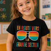 Jeune fille souriante portant un t-shirt noir avec le design « En route vers la grande section » dans une salle de classe d'école maternelle.