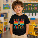 Jeune enfant souriant portant un t-shirt noir avec le design « En route vers la moyenne section » dans une salle de classe d'école maternelle.