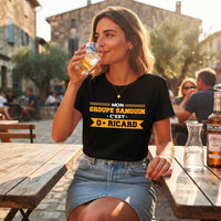 Une femme assise à une table de café en terrasse en Provence, portant le t-shirt noir avec le design jaune "MON GROUPE SANGUIN • C'EST • O+ RICARD". Elle boit dans un verre de Ricard avec de la glace. Une bouteille de Ricard et un pichet d'eau sont sur la table en bois. L'arrière-plan est flou avec des bâtiments en pierre et d'autres clients.