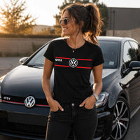 Femme portant un t-shirt noir avec un design GTI et le logo Volkswagen sur la poitrine, debout devant une Volkswagen Golf GTI noire dans un décor urbain au coucher du soleil.