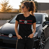Femme portant un t-shirt noir avec un design GTI et le logo Volkswagen sur la poitrine, debout devant une Volkswagen Golf GTI noire dans un décor urbain au coucher du soleil.