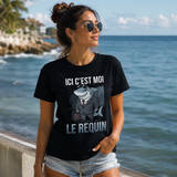 Femme portant un t-shirt noir avec le design « Ici c'est moi le requin » devant la mer sur une promenade en bord de l'eau