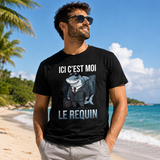 Homme portant un t-shirt noir avec le texte « ICI C'EST MOI LE REQUIN » et une illustration d'un requin en costume, photographié sur une plage tropicale avec le sable, les palmiers et l'océan en arrière-plan.
