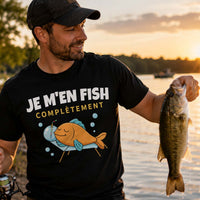 Homme portant un t-shirt noir avec l'inscription « Je m'en Fish Complètement », tenant une canne à pêche et un poisson fraîchement pêché au bord d'un lac au coucher du soleil, dans une ambiance paisible et naturelle.