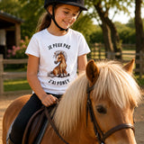 Fille portant un t-shirt blanc avec l'inscription « Je peux pas J'ai Poney » et une illustration de poney, montée sur un poney dans un centre équestre en plein air avec une carrière, des clôtures en bois et des arbres en arrière-plan.