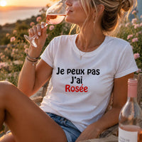 Femme portant un t-shirt blanc avec l'inscription « Je peux pas J'ai Rosée », dégustant un verre de vin rosé sur une terrasse en fin de journée, avec une bouteille de rosé, des fleurs et un paysage ensoleillé en arrière-plan.
