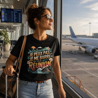 Femme portant un t-shirt noir avec le design « Je peux pas, je me barre à La Réunion » dans un terminal d'aéroport avec une valise devant les baies vitrées donnant sur les avions