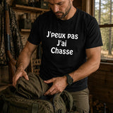 Homme portant un t-shirt noir avec l'inscription « J'peux pas J'ai Chasse », préparant son sac et son équipement de chasse dans un chalet en bois avant de partir en forêt.