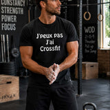Homme portant un t-shirt noir avec l'inscription « J'peux pas J'ai Crossfit », se préparant à un entraînement dans une salle de CrossFit équipée de cages de musculation, d'anneaux de gymnastique, de box en bois et d'une kettlebell.