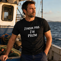 Homme portant un t-shirt noir avec le design « J'peux pas J'ai Pêche » debout sur un bateau de pêche en mer, entouré de filets et regardant l'horizon.