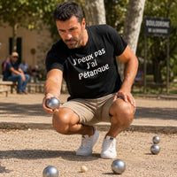 Homme portant un t-shirt noir avec le design « J'peux pas J'ai Pétanque » en train de viser le cochonnet lors d'une partie de pétanque sur un boulodrome ensoleillé.