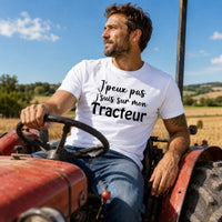 Homme portant un t-shirt blanc avec le design « J'peux pas j'suis sur mon Tracteur » au volant d'un tracteur dans un champ agricole en pleine campagne.