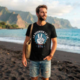 Un homme barbu porte le t-shirt noir imprimé du design "LA RÉUNION LE LA" avec une main faisant le signe de la paix. Il porte un short en jean déchiré, une montre et un sac en paille à l'épaule. Il est debout sur une plage de sable noir volcanique avec des montagnes verdoyantes en arrière-plan sous une lumière de fin de journée.
