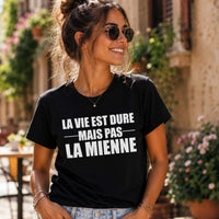 Femme portant un t-shirt noir avec l'inscription « La vie est dure mais pas la mienne », souriante dans une rue pittoresque bordée de fleurs, avec des façades en pierre et des terrasses de café en arrière-plan.