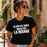 Femme portant un t-shirt noir avec l'inscription « La vie est dure mais pas la mienne », souriante dans une rue pittoresque bordée de fleurs, avec des façades en pierre et des terrasses de café en arrière-plan.