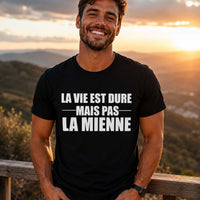 Homme portant un t-shirt noir avec l'inscription « La vie est dure mais pas la mienne », souriant face à un panorama de montagnes au coucher du soleil, dans une ambiance positive et inspirante.