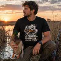 Homme portant un t-shirt noir avec une illustration de canard et le texte « Le canard m'appelle, il faut que j'y aille », photographié dans un marais au coucher du soleil en tenant un canard colvert, avec les roseaux et un plan d'eau en arrière-plan.