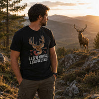 Homme portant un t-shirt noir avec une illustration de tête de cerf et le texte « Le cerf m'appelle, il faut que j'y aille. », photographié dans un paysage de montagne au coucher du soleil, observant un grand cerf au loin sur un promontoire rocheux.