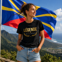 Femme portant un t-shirt noir avec l'inscription dorée « LÉGENDE NÉE À LA RÉUNION », photographiée devant le drapeau de l'île de La Réunion avec un paysage montagneux verdoyant en arrière-plan. 