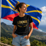 Femme portant un t-shirt noir avec l'inscription dorée « LÉGENDE NÉE À LA RÉUNION », photographiée devant le drapeau de l'île de La Réunion avec un paysage montagneux verdoyant en arrière-plan. 