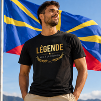 Homme portant un t-shirt noir avec l'inscription dorée « LÉGENDE NÉE À LA RÉUNION », photographié devant le drapeau de l'île de La Réunion dans un paysage montagneux ensoleillé.