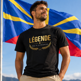 Homme portant un t-shirt noir avec l'inscription dorée « LÉGENDE NÉE À LA RÉUNION », photographié devant le drapeau de l'île de La Réunion dans un paysage montagneux ensoleillé.
