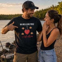 Pêcheur portant un t-shirt noir avec le design « Ma Meilleure Prise, c'est elle » en montrant sa compagne du doigt au bord d'un lac lors d'une sortie de pêche en couple.