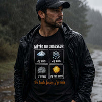 Homme portant un t-shirt noir avec l'inscription « Météo du Chasseur », marchant sous une pluie battante sur un chemin forestier, équipé d'une veste imperméable, dans une ambiance de chasse automnale.