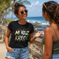 Jeune femme souriante portant un t-shirt noir avec le design « Mi Koz Kréol » en train de discuter au bord d'une plage tropicale sous les palmiers.