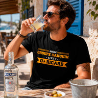 Homme portant un t-shirt noir avec le design « Mon groupe sanguin c'est O+ Ricard » en train de boire un pastis sur la terrasse ensoleillée d'un café méditerranéen.