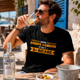 Homme portant un t-shirt noir avec le design « Mon groupe sanguin c'est O+ Ricard » en train de boire un pastis sur la terrasse ensoleillée d'un café méditerranéen.