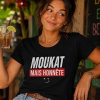 Femme au sourire malicieux portant un t-shirt noir avec le design « Moukat mais honnête » dans un bar tropical en plein air, un verre à la main.