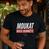Homme au sourire malicieux portant un t-shirt noir avec le design « Moukat mais honnête » dans un bar créole tropical.