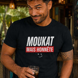 Homme au sourire malicieux portant un t-shirt noir avec le design « Moukat mais honnête » dans un bar créole tropical.