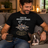 Homme portant un t-shirt noir avec le design « Papa d'un Bouledogue Français – Partenaire officiel de sieste » assis dans un salon confortable avec son Bouledogue Français endormi à ses côtés.