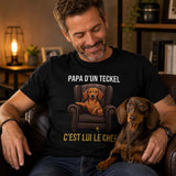 Homme portant un t-shirt noir avec le design « Papa d'un Teckel, c'est lui le chef » assis dans un salon chaleureux avec son chien Teckel sur les genoux.